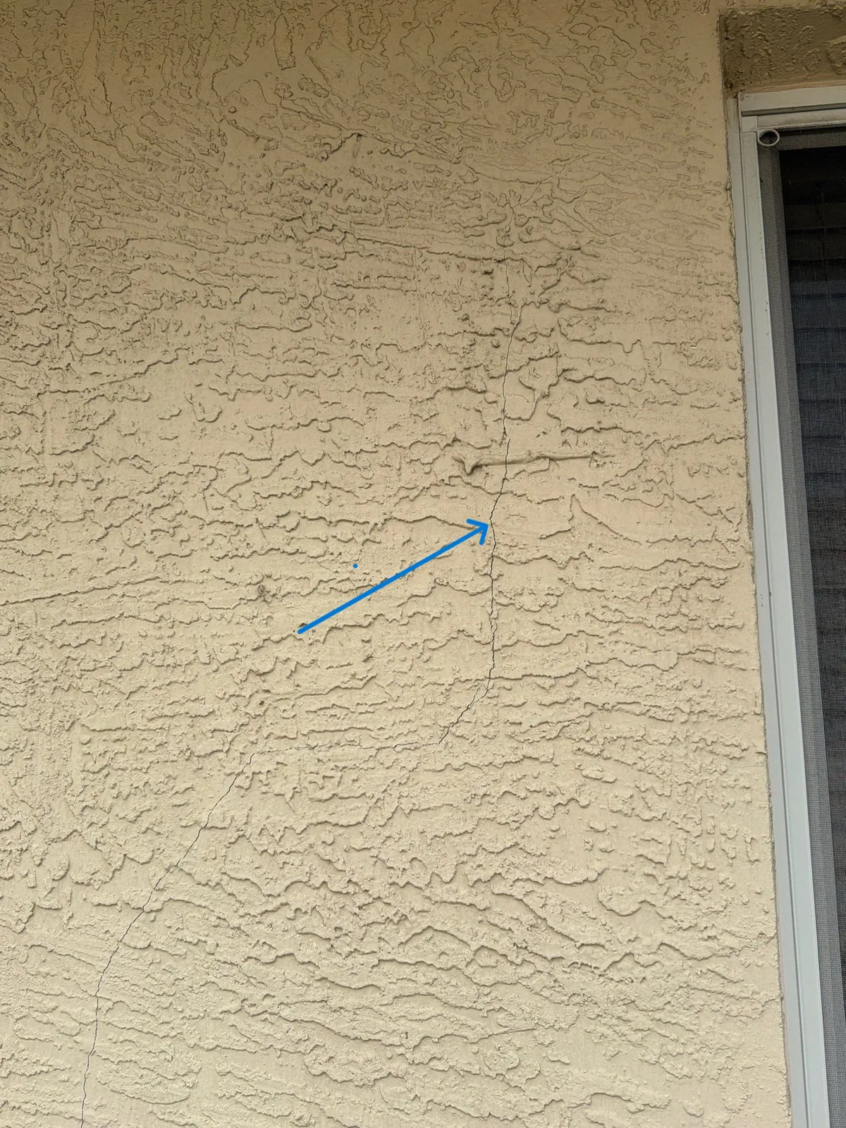 Hairline crack running vertically through textured stucco, marked with an arrow before repair
