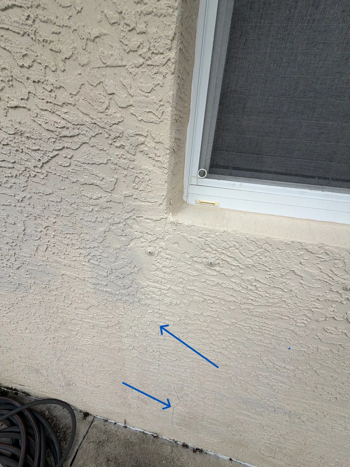 Hairline cracks in stucco below a window corner and at the base of the wall, marked with arrows before repair